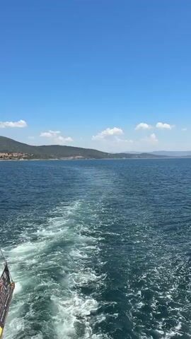 The video captures a serene daytime scene from a moving boat on the water in Menderes, Turkey. Initially, the camera is positioned at the stern, looking back at the boat's churning white wake as it cuts through the deep blue sea. In the background, a distant coastline featuring green and brown hills or mountains stretches across the horizon under a vast, clear blue sky dotted with fluffy white clouds.

As the video progresses, the camera slowly pans forward, revealing the open expanse of the sea. The boat's wake gradually moves out of the frame, showcasing the calm, slightly rippled surface of the water leading towards the distant landmass. The bright sunlight suggests a clear day, possibly late morning or early afternoon. A small, indistinct part of the boat with some unreadable text or graphics is briefly visible on the bottom left edge at the very beginning of the clip.