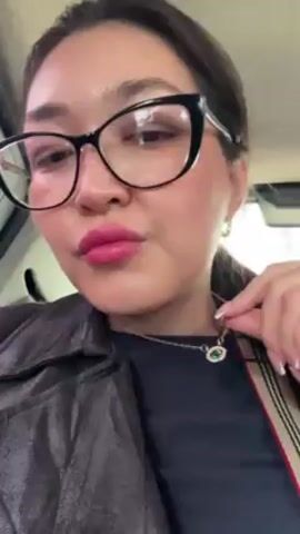 A woman with dark hair, wearing black glasses and pink lipstick, is indoors, possibly in a car. She is wearing a black top and a brown jacket.  She holds up a gold necklace with a green pendant and then puts it on.