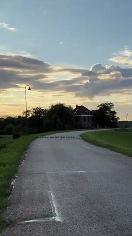 A vertical video shows a paved road curving through a grassy landscape under a cloudy sky. A two-story brick house with a dark roof sits nestled among trees on the left side of the road. A street lamp stands near the road on the left. Further down the road on the right, a small building with orange and beige sections is visible. The grass is green and lush, suggesting a rural setting. The sky is a mix of blue and white clouds, with hints of orange and yellow light, possibly indicating sunset or sunrise. The road appears to be slightly wet in places. There are no people visible in the video.