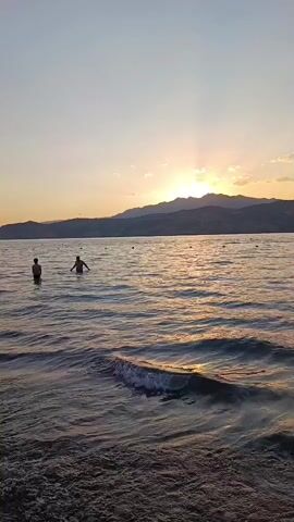 The video shows a body of water with waves, a shoreline, and mountains in the background. Several people are in the water. The sun is setting, casting a warm glow over the scene.