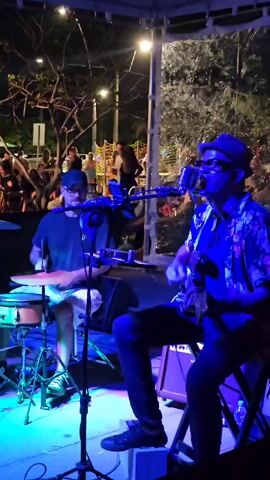Two musicians perform live under a gazebo for a small crowd at night. The musician on the right plays guitar and sings into a microphone, wearing a floral shirt, black pants, a fedora, and glasses. The musician on the left plays drums, wearing a black t-shirt, white shorts, and a baseball cap. The stage is lit with blue and purple lights. The audience is a mix of people, some standing, some walking, some dancing. Trees are visible in the background.