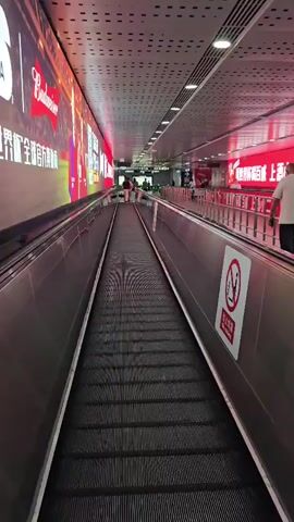 A first-person perspective video captures movement along a moving walkway in a brightly lit, modern hallway. The walkway is bordered by stainless steel railings. On the left, a large, vibrant digital billboard displays advertisements, primarily featuring the Budweiser logo and images of soccer players. On the right, a similar digital billboard is visible, also illuminated with red tones and featuring text in Chinese characters. Several people are seen walking in both directions on the moving walkway and in the distance. A red and white sign with a no smoking symbol and Chinese characters is affixed to the railing on the right. The ceiling is perforated with a grid of circular lights. The overall impression is a high-traffic, commercial environment, possibly an airport, train station, or shopping mall.