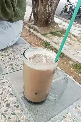 The video opens with a close-up shot of a tall glass mug filled with a brown, iced beverage and a green straw. The mug has a handle. The glass sits on a textured surface that appears to be a table or bench with a speckled pattern. In the background, a person's legs, clad in light-colored pants and a green top, are visible on the left side of the frame. The background also shows a large tree trunk, a sidewalk or pathway with scattered leaves and debris, and beyond that, a paved area where several motorcycles are parked. The camera then pans slightly to the right and upwards, revealing more of the motorcycles and the surrounding environment, which seems to be an outdoor public space. The lighting suggests it is daytime. The video is short and seems to be a casual observation of a drink and its surroundings.