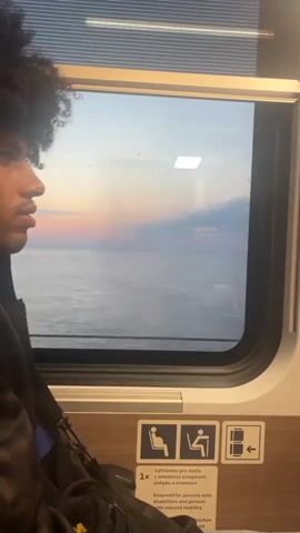 The video shows a young person with curly dark hair looking out a train window. The person is wearing a dark jacket over a blue shirt and has a black bag with yellow accents on their lap. The train window reveals a wide expanse of calm water, possibly the sea, under a pale sky with streaks of pink and orange from a sunset. There are also some white clouds visible. Below the window, there are three signs. The leftmost sign depicts a seated person, indicating a seat. The middle sign shows a person seated at a table with a laptop, signifying a table seat. The rightmost sign illustrates a person in a wheelchair with an arrow pointing to the right, indicating an accessible space. A smaller sign below the leftmost two states "Reserved for persons with disabilities and persons with reduced mobility" in English and likely another language. The train interior is visible in the foreground, with a textured headrest area. The lighting suggests it is dusk or dawn.