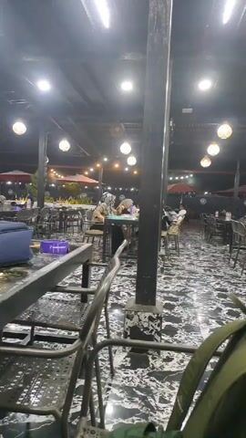 The video captures an indoor or semi-outdoor restaurant or cafe at night, illuminated by various artificial lights. Numerous dark-colored metal tables and chairs are arranged across a floor with a distinctive black and white marbled pattern. Several patrons are seated at different tables throughout the establishment, engaging in casual activity such as dining or socializing. Two individuals are more clearly visible in the mid-ground, seated at a table; one person wears a headscarf. In the background, more seating areas are visible, along with large red umbrellas, suggesting an outdoor dining section. Bright linear lights and warm, spherical hanging lamps provide ample illumination. A blue bag and a purple container rest on a table in the foreground. Part of a chair and a backpack are visible in the bottom right, close to the camera. The scene conveys a relaxed and social atmosphere. No discernible text is visible in the video.