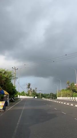 The video begins with a shot of a road under a cloudy sky. The road is wet, suggesting recent rain. There are power lines with several birds perched on them. Palm trees and other greenery line the sides of the road. A white barrier with black and white markings separates the road from the sidewalk. As the video progresses, the perspective shifts to a selfie video taken by a person wearing a red helmet and sunglasses. The person appears to be riding a motorcycle or scooter, as their bare shoulders are visible and the wind is blowing their hair. The background shows more trees and what appear to be small roadside stalls. The sky is still cloudy in the background, but brighter.