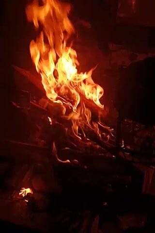 The video shows a fire burning intensely. Flames rise and dance around partially visible wooden structures within the fire. The scene is dark, with the fire providing the main source of illumination.