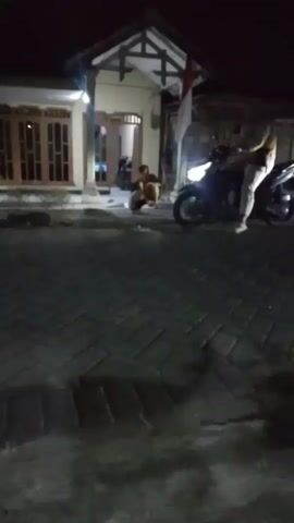At night, a person is crouched on a paved driveway in front of a house. A motorcycle is parked nearby with its headlight on. Another person is standing near the motorcycle. The house has a covered porch with a flag displayed. The scene is dimly lit.