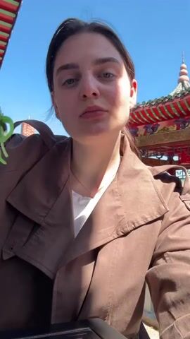 A young woman with dark hair pulled back is captured in a close-up, outdoors on a sunny day. She is wearing a brown trench coat over a white t-shirt and a delicate gold necklace. In her right hand, she holds a pair of wooden chopsticks, lifting a portion of green, leafy food (likely seaweed salad) towards her mouth.

The video shows her in two main actions: initially, she looks forward with the food poised, and then she tilts her head back to put the food into her mouth. Her expression is focused as she eats.

The background features a clear, bright blue sky. Behind her, parts of an ornate structure with a green tiered roof and red decorative elements, resembling a pagoda or temple-like building, are visible. This architectural style suggests an East Asian-themed setting. The bright sunlight indicates it is daytime, likely mid-day, with no clouds in sight. The overall impression is one of enjoying a meal outdoors in a pleasant, culturally inspired environment, possibly in Hainburg an der Donau, Austria, as per the provided location context.