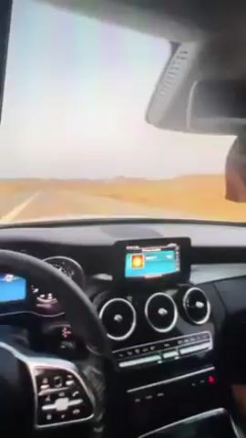 Okay, here's a concise description of the video:

The video is shot from inside a car, seemingly a Mercedes-Benz based on the interior design. The car is driving on a straight road through a desert landscape with a pale, almost white sky.  The camera angles shift, showing the dashboard with the infotainment screen, the air vents, and the steering wheel. There's also a brief shot of a passenger in the back seat. The driver is visible periodically, steering and interacting with the car. Music is playing in the background. The speedometer is visible briefly, indicating speed.