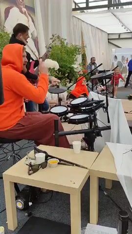 A group of musicians perform outdoors. In the foreground, a man wearing an orange hooded sweatshirt and brown pants plays an electronic drum kit. Next to him, a man in a white t-shirt plays an acoustic guitar. Behind them, another musician plays a red electric bass guitar. A woman in an orange headscarf can be seen in the background playing a guitar. The musicians are set up under a white canopy, with greenery and large floral arrangements visible behind them. In the foreground, a small table holds sunglasses, cups, and a small candle. The lighting suggests it is daytime.