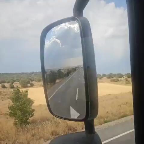 The video is a first-person perspective shot from inside a vehicle, looking at the side-view mirror. The mirror reflects a scene of a rural landscape with a road stretching into the distance. The sky is partly cloudy with sunlight breaking through. The road appears to be asphalt and has a white line marking the lanes. On either side of the road, there are fields of dry grass and scattered trees. The mirror itself is black and rectangular, mounted on a black arm. There are no people or other vehicles clearly visible in the reflection, though distant shapes on the road might be vehicles. The overall impression is one of travel through an open, sunny landscape.