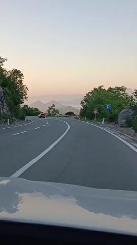 The video shows a view from the windshield of a car driving on a winding road. The road has two lanes, separated by a white line. There are white dashed lines on the right side of the road. On the left side of the road, there are trees and rocks. On the right side of the road, there are trees, rocks, and guardrails. There are also road signs on the right side of the road: a yellow triangular sign with a red border and a black arrow indicating a curve, and a blue square sign with a white letter P indicating parking. In the distance, there are mountains and a body of water, possibly a lake or sea, under a pale sky with a hint of orange from the sunrise or sunset. A red car is visible ahead on the left lane of the road. The car's hood is visible in the foreground, reflecting the sky.