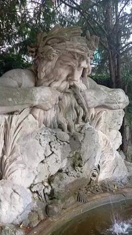 This is a close-up shot of a large, weathered stone statue of a man with a long beard and a laurel wreath on his head. He appears to be reclining or resting, with his arms bent and his head tilted downwards. The statue is situated outdoors, with lush green foliage and trees visible in the background. The statue is partially submerged in a pool of water, which appears to be part of a fountain, as a stream of water is visible splashing into the pool from the right side of the frame. The stone of the statue is rough and textured, with some patches of moss or algae growing on its surface, suggesting it has been exposed to the elements for a significant period. The lighting suggests it is daytime, with diffused light filtering through the trees. The overall atmosphere is serene and tranquil, evoking a sense of timelessness and natural beauty. The location is identified as Baden, Austria.
