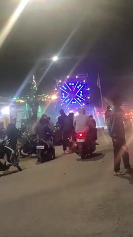 A group of people are gathered on motorcycles at night, facing a stage with bright lights and a large speaker setup. The stage is illuminated with purple and blue lights, and spotlights are visible. One person in the foreground, standing on the right side of the road, is holding a red signaling stick. Vehicles, including motorcycles and cars, are parked and moving in the background. The environment appears to be an outdoor event, possibly a concert or festival, with a road or open space in front of the stage.