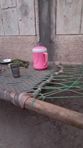 The video shows an outdoor scene with a pink insulated mug and a metal cup on a woven mat. There is a bundle of green herbs or plants next to the cup and some small objects that appear to be nuts or seeds on a metal plate to the left. The mat is made of fabric strips and is stretched over a wooden frame, typical of a charpai or cot. The background consists of a textured wall with a vertical concrete pillar or seam. The lighting suggests it is daytime, possibly late afternoon or early morning, given the shadows and soft light. The overall environment appears to be humble, possibly rural or in a less developed area. There are no people visible in the video. The camera is positioned at a low angle, focusing on the objects on the mat.