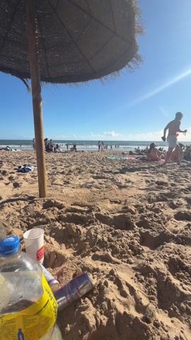The video captures a sunny beach scene with a clear blue sky and scattered clouds. People are visible in the distance, some swimming or standing in the water, while others are sunbathing or socializing on the sand. The foreground is dominated by sand, with a thatched beach umbrella providing shade in the upper left corner. Litter is scattered on the sand, including a plastic bottle of juice, a paper cup, and two empty energy drink cans. The overall impression is one of a crowded beach on a bright, sunny day, with signs of leisure activities and discarded waste.