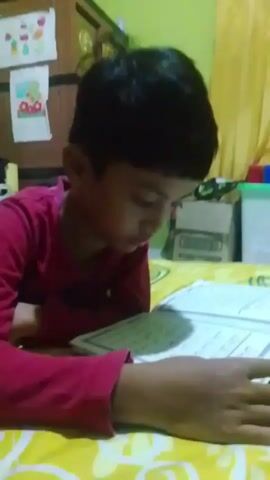 A young boy wearing a red shirt is lying on a yellow and white patterned surface and reading a book with Arabic text. He is looking down at the book, his right hand resting on the pages. The background is blurred, but appears to be a room with some objects on shelves. The lighting is dim, suggesting it may be indoors or nighttime.