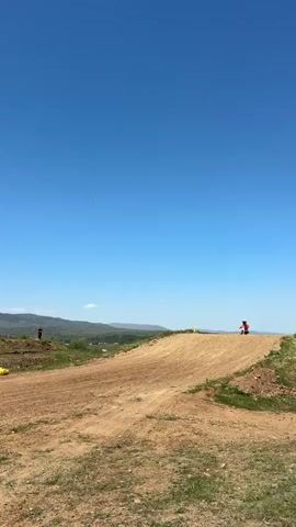 The video shows a motocross race on a sunny day. A child on a small motorcycle is riding along a dirt track, with other people standing around watching. There are yellow barriers on the track and in the background a landscape of hills and trees. Some people in the foreground are wearing racing credentials around their necks. The sky is clear and blue.