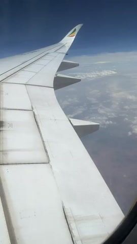 The video shows the wing of an Ethiopian Airlines plane as seen through the window during flight. The wing is white, and the Ethiopian Airlines logo, a green, yellow, and red flag, is visible on the wingtip. Below the plane, there are clouds and land. The sky is blue. The camera slightly pans across the wing.