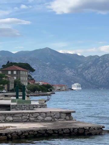 The video shows a scenic view of the water, mountains, and buildings in Kotor, Montenegro. The water is blue and has small waves. There is a large cruise ship visible on the water in the distance. There is a dock in the foreground with green posts. A dark green SUV with a roof rack drives in front of the camera during the video. The sky is blue with some white clouds. The mountains are rocky and have some vegetation. The buildings are old and have red tile roofs. The video appears to have been shot on a clear, sunny day.