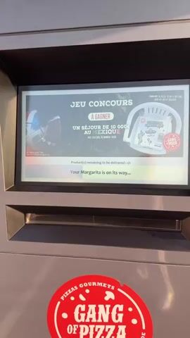A close-up shot focuses on a touchscreen display mounted on a grey outdoor kiosk. The screen shows a promotion for a contest, "JEU CONCOURS," with a grand prize of a 10,000 Euro trip to Mexico. Below the contest details, a message reads "Product(s) remaining to be delivered: 1/1" followed by "Your Margarita is on its way...". The bottom of the kiosk features a red circular sticker that says "PIZZAS GOURMETS GANG OF PIZZA". The camera then pans slightly to the left, revealing more of the kiosk's exterior, which is grey and appears to be made of metal. To the left of the kiosk, a brick wall is visible, plastered with promotional posters for pizzas. One poster advertises "PIZZAS EXECUTED IN 3 MINUTES" and another details "UN SAVOIR-FAIRE RECONNU" (Recognized know-how) with images of pizza preparation. Next to the kiosk, black trash bags are piled up. The background suggests an outdoor setting with some foliage and a railing visible. The lighting indicates it is daytime.