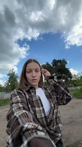 A young woman is holding a camera and taking a selfie video on a dirt road. She is wearing a white t-shirt and a brown plaid flannel shirt. She is outdoors in a rural area. There are trees, a blue sky with white clouds, and a power line pole nearby. A red truck passes by, and a small child walks in the background. The camera is rotated at varying angles throughout the video.