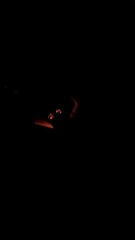The video begins with a dark scene, showing two blurry red lights in the distance, possibly car taillights. The camera then pans to the right, revealing a dark interior of a vehicle. The dashboard is visible, with a glowing blue display and various red illuminated buttons. A speedometer is also partially visible, with a needle pointing to a speed. The scene suggests the video was taken at night, possibly while driving.