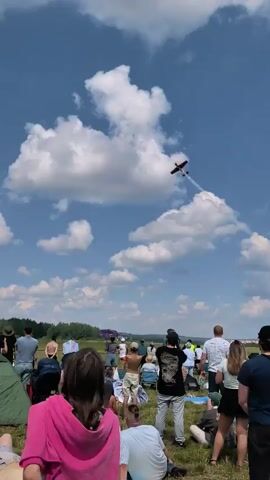This is a low-angle, vertical video of an outdoor event on a sunny day with scattered clouds. A small, red propeller plane is performing aerial maneuvers in a blue sky. The plane is leaving a white smoke trail as it flies and executes a turn. Below the plane, a crowd of people is gathered, facing upwards to watch the display. Some individuals are holding up cell phones to record the event. In the foreground, tents and green foliage are visible, indicating a field or park setting. The overall atmosphere appears to be one of enjoyment and shared spectacle.