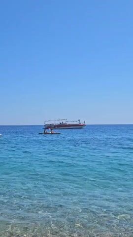 The video opens with a wide shot of a bright blue sky and calm, clear turquoise water. In the distance, a red and white passenger boat with an awning is visible. Closer to shore, two people are in a kayak. The camera then pans down to a pebble beach, where a blue sarong is spread out, and a black portable speaker lies on top of it. The sound of waves can be heard. The footage is shot from a low angle, looking out towards the water.