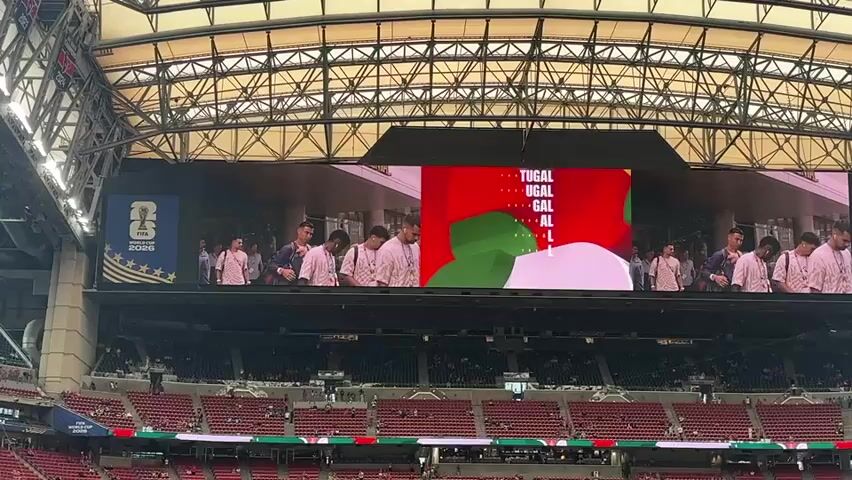 This media file captures a view of a large stadium screen displaying the Portugal national team's participation in the FIFA World Cup 2026. The screen prominently features the word "PORTUGAL" in large white letters against a red background, flanked by the Portuguese flag on either side. Below the text, a stylized representation of the Portuguese flag's green and red colors is visible.

On the screen, several individuals, presumably members of the Portuguese football team, are shown walking towards the camera. They are dressed in casual attire, with some wearing light-colored shirts. The background on the screen appears to be an outdoor setting with modern architecture.

To the left and right of the main screen, two identical panels display the FIFA World Cup 2026 logo, emphasizing the event. The stadium itself is filled with rows of red seats, many of which are occupied by spectators, though they appear as distant figures. The overall lighting suggests it is daytime, and the atmosphere is likely that of a pre-match event or broadcast. The scene is set within a large, modern stadium structure with a visible metallic roof framework. The presence of the FIFA World Cup branding and the national team's display indicates a significant sporting event.