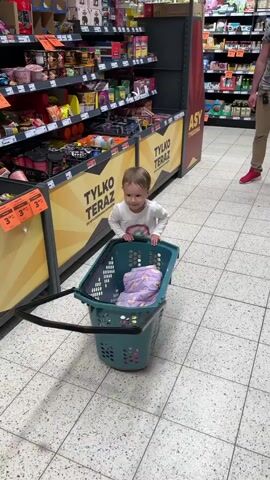 In a brightly lit supermarket, a young child stands in an aisle with a turquoise shopping basket in front of them. The basket contains a folded purple patterned garment. The child, wearing a white long-sleeved shirt with a rainbow design and pink pants and shoes, is smiling and clapping. The child then walks around and runs to the right. Supermarket shelves line the aisle with various products. Signage on the shelf displays the words "TYLKO TERAZ" in yellow letters. An adult with red shoes is walking in the background. The floor is tiled in light gray and white.
