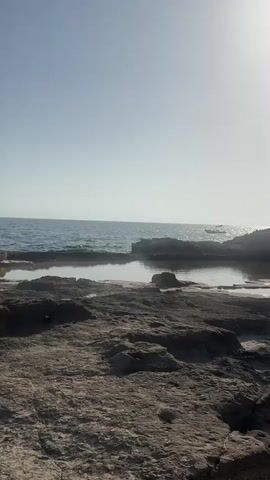 The video shows a calm ocean scene with a rocky shoreline in the foreground. The sun is shining brightly, casting a gentle glare on the water. The water is a deep blue, and the sky is a clear, pale blue. There are a few rocks scattered in the water, forming small pools. A small white boat can be seen in the distance on the right side of the frame. The overall mood is peaceful and serene.