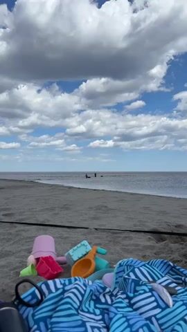 The video shows a beach scene on a partly cloudy day. In the foreground, there are colorful children's beach toys and a blue patterned beach blanket on the sand. In the distance, two people are standing in the water, each holding a long pole and seemingly working with what look like boxes or containers in the water. The beach stretches across the middle ground, leading to the water and the two people. The sky is filled with fluffy white clouds, with patches of blue sky visible between them.