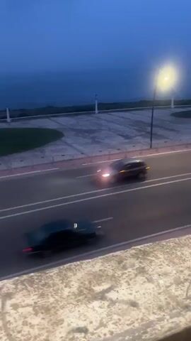 The video shows a gray car driving on a multi-lane road next to a grassy area and the ocean. The sky is a deep blue, suggesting it is dusk or early morning. The camera appears to be positioned at a high angle, overlooking the scene. The car is the primary object in motion. The lighting is dim and mostly natural.