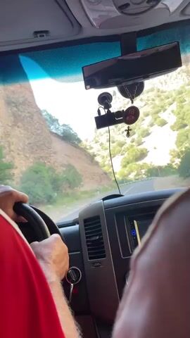 The video shows the interior of a vehicle from the passenger's perspective. A person wearing a red shirt is driving, their hands visible on the steering wheel. The dashboard features a car stereo, air vents, and a key in the ignition. Several objects are attached to the windshield via suction cups, including a dash camera, a rearview mirror, and religious icons. The road ahead curves through a mountainous landscape with steep, rocky slopes covered in sparse vegetation. The sky is clear and bright.
