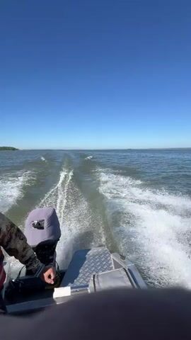 A first-person perspective video shows the rear of a boat moving through water. The boat has a grey outboard motor with a black cover on top, and a person wearing a camouflage jacket and a life vest is partially visible on the left side. The boat is creating a large wake, indicated by white foamy trails in the water behind it. The sky is clear and blue, and the horizon is visible in the distance with a faint line of land. The water appears calm, with small ripples on the surface. The video is shot from a stationary position within the boat, looking backward.