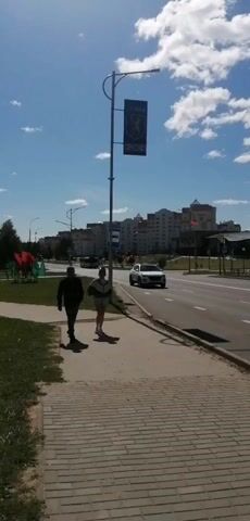 A wide shot shows a paved sidewalk with two people walking away from the camera. The person on the left is wearing a dark jacket and pants. The person on the right is wearing a white jacket with dark stripes and light shorts. They are walking on a sunny day with a blue sky and scattered clouds. In the background, there is a street with cars, a lamppost with a banner, and buildings.