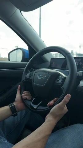 A person is sitting in the driver’s seat of a car, holding the steering wheel with both hands. The car’s infotainment display screen is visible in the background. The steering wheel features the logo of a running horse, indicating the car is a Ford Mustang. The driver is wearing a watch on their left wrist and blue jeans. The car is moving, as the scenery outside the windshield is changing.  Music is playing in the background.