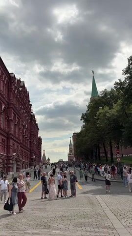 The video shows a wide cobblestone street lined with people. On the left is a large, ornate red building. On the right, trees form a boundary with a tall green spire visible behind them, and further down the street, a shorter tower with a clock is visible. Numerous people are walking or standing along the street, many of them appear to be tourists. Some individuals are in the foreground, facing away from the camera, while others are further down the street. The sky is cloudy.