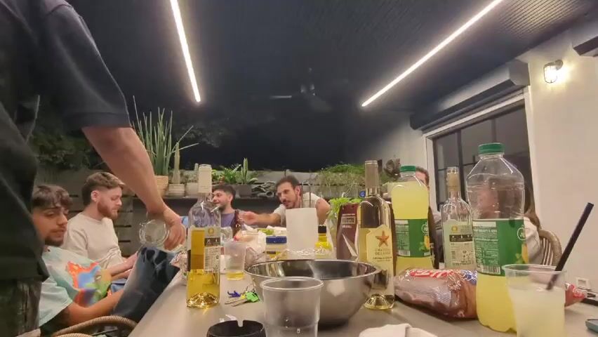 A group of people are gathered around a table on an outdoor patio at night. One person, whose body is partially in the frame, is pouring a liquid from a clear glass bottle into a glass held by someone seated. On the table are bottles of alcohol, lemons, lemonade, condiments, a metal bowl, and other items. The patio is covered with a dark ceiling and lit by multiple fluorescent lights. A window with a roll-up shade is visible in the background, along with plants in pots. People are talking and interacting with each other.
