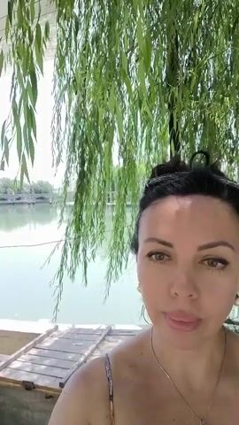 A woman with dark hair pulled back and wearing a patterned sundress is filming a selfie video outdoors. She is positioned in the foreground, with a body of water and trees behind her. The water appears calm, reflecting the sky and foliage. A wooden platform or dock is visible in front of her. In the background, there are some structures that look like outdoor seating or gazebos. There are also some garden gnomes and greenery. The overall atmosphere is relaxed and suggests a warm, sunny day in a natural setting, possibly by a lake or river.