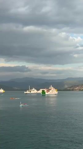 A wide shot shows a body of water with several boats and ships. In the foreground, a small blue and red boat with a person on it is moving left to right. Further back, a few yellow boats are also moving left to right. Two large white ships with red accents are visible, one closer to the right and another further left. Both ships are stationary or moving slowly. The water is a deep blue-green, and the sky is filled with dramatic, dark grey clouds, with patches of lighter sky and sunlight breaking through. In the background, a range of blue-green mountains stretches across the horizon. The overall impression is a calm, yet somewhat moody, maritime scene.