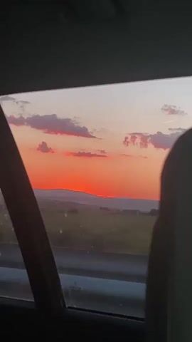 This video captures a view through a car window during a sunset. The initial shot shows a dark interior of a car and a hazy exterior. As the video progresses, the sky transitions to a vibrant sunset with hues of orange and pink, illuminating scattered clouds. A rural landscape with rolling hills and fields is visible below the sky, and power lines run horizontally across the frame. The sun is low on the horizon, partially obscured by a distant hill. The perspective suggests the viewer is a passenger in the moving vehicle, looking out the rear window.