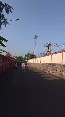 The video shows a straight asphalt road lined with a red wall on the left and a tall, cream-colored wall with a stone foundation on the right. Several people are walking along the road. On the right side of the road, a tall stadium light pole is visible, along with a fenced structure behind the wall. The sky is clear and blue, suggesting it is daytime.