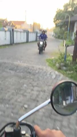 The video shows a first-person perspective of someone riding a motorcycle on a paved road. In the distance, another person is riding a motorcycle towards the camera. The environment appears to be a residential area with houses and fences on either side of the road. The lighting suggests it is either early morning or late afternoon. The camera is slightly shaky, indicating movement. The focus is primarily on the road ahead and the approaching rider.