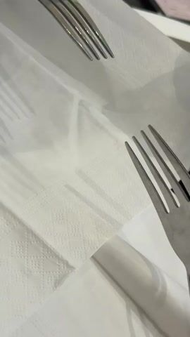 Two metal forks are positioned on white paper napkins. The fork on the left is partially out of focus, and its tines show iridescent coloring, suggesting heat or a chemical reaction. The fork on the right is in focus, and its tines also exhibit similar iridescent colors. A shadow of the fork on the left is visible on the napkins. A hand is partially visible in the lower right corner of the frame.