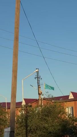 A concrete utility pole is in the foreground of the video, with telephone wires stretching across the frame. To the right of the pole, a flag of Kazakhstan is visible, attached to a smaller pole. Behind the flag and utility pole, a brick building with a red roof can be seen, partially obscured by green foliage. The sky is clear and blue. A street lamp extends from the utility pole to the left. Another street lamp is visible in the distance.
