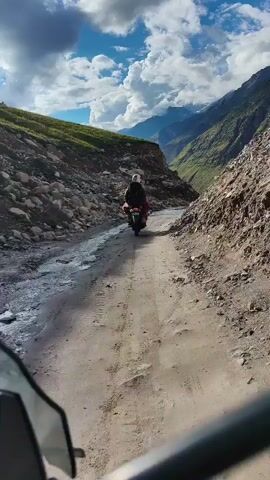 The video shows a motorcycle with a rider from a first-person perspective. The motorcycle is traveling on a dirt road that is rough and uneven, with rocks and debris scattered across it. The road is situated between steep, rocky mountainsides covered in greenery. The sky above is partly cloudy, with dramatic white clouds against a blue sky. The rider is wearing a helmet and a backpack, and the motorcycle appears to be loaded with luggage, suggesting a journey or adventure. The camera is mounted on another motorcycle, following the lead rider. The scene suggests a remote and challenging terrain, possibly in a mountainous region during the daytime.