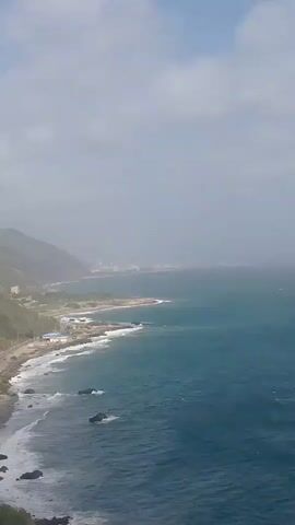 This is a video of a coastal landscape. The scene looks out over a wide expanse of the ocean. In the foreground, waves crash against a rocky shore. The water is a deep blue-green, with white foam from the breaking waves. Several dark rocks are visible in the water near the shore.

Further along the coast, there is a stretch of beach with some buildings. A road runs parallel to the coastline, with some traffic visible on it. Mountains rise up from the land behind the road, covered in green vegetation. In the distance, a hazy city skyline can be seen along the coast, suggesting a populated area. The sky is overcast with scattered clouds, and the overall atmosphere appears to be somewhat muted due to the weather. The time of day appears to be daytime, likely morning or afternoon, given the lighting. There is no visible text in the video.